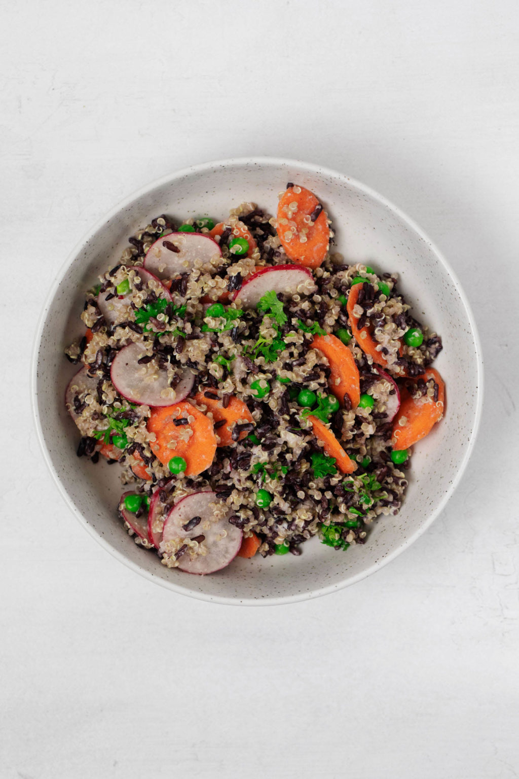 A plant-based grain salad with carrots and peas is served with herbs in a white, round serving bowl.