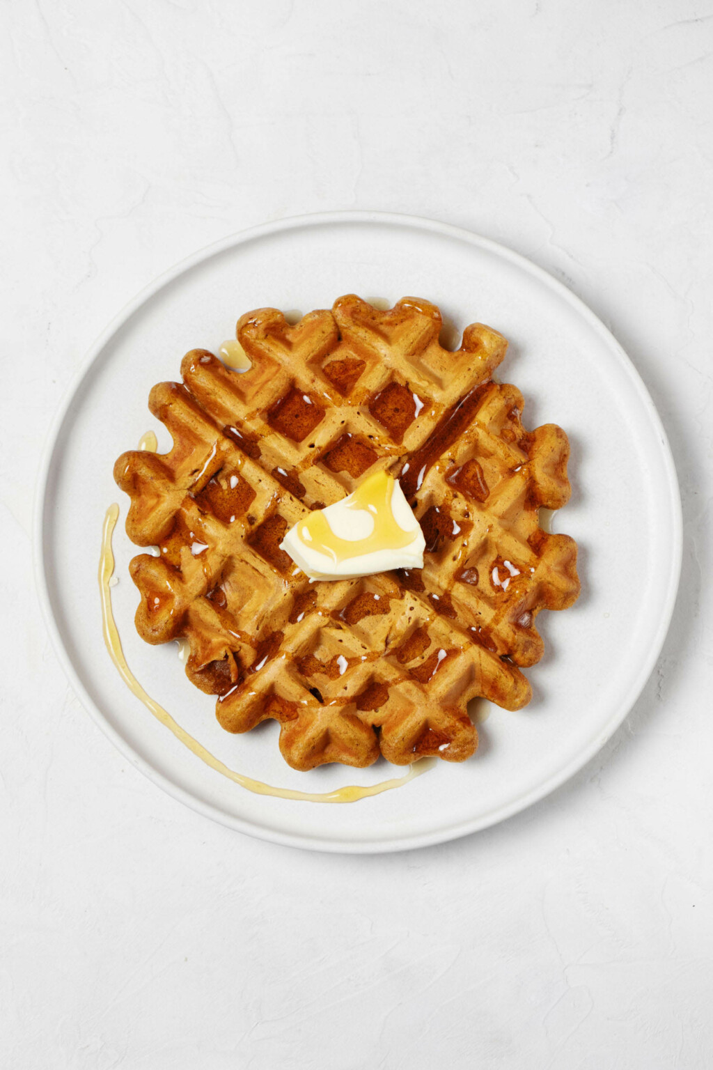 A round, white plate rests on a white surface. The plate is being used to serve a vegan pumpkin waffle, which is topped with plant-based butter and maple syrup.