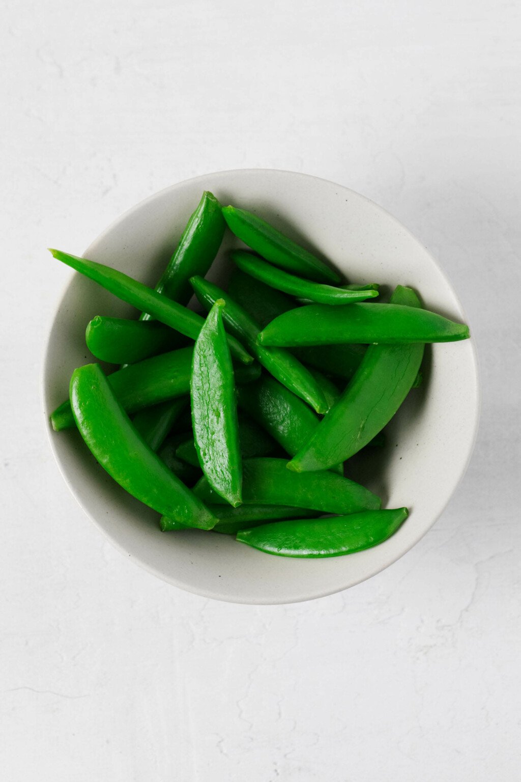 An overhead image of snap peas in a small, round white bowl.