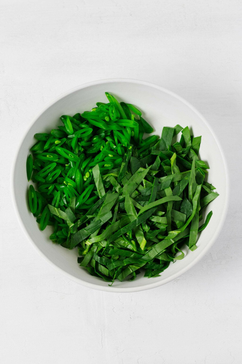 An overhead image of a mixing bowl, which contains thinly sliced snap peas and baby spinach leaves.
