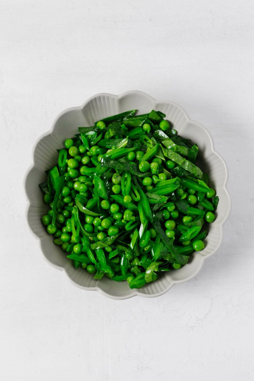 A round, fluted white bowl is filled with bright green, fresh peas and thinly sliced snap peas, along with spinach. The vegetables glisten lightly with a vinaigrette.