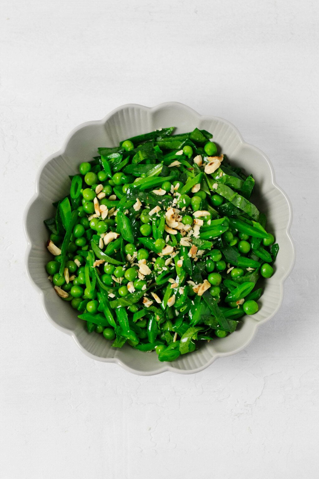 A round, fluted white bowl is filled with bright green, fresh peas and thinly sliced snap peas, along with spinach. The vegetables glisten lightly with a vinaigrette. There are finely chopped roasted nuts on top.
